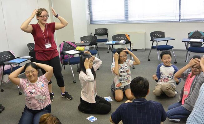 A teacher stands while a group of elementary school students sit in a circle on the floor playing a game