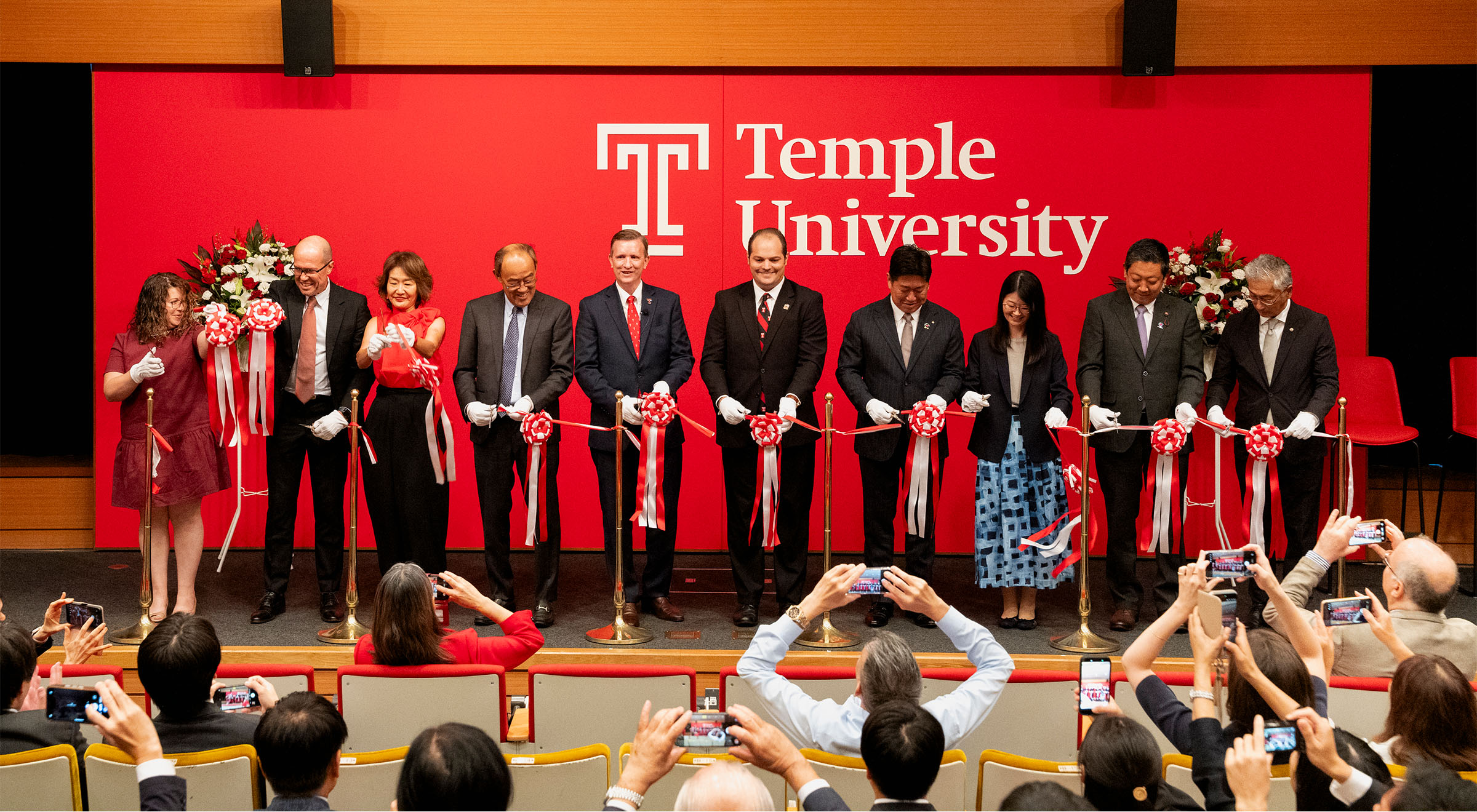TUJ leaders and distinguished guests cut a ribbon on stage in front of a red Temple University backdrop.